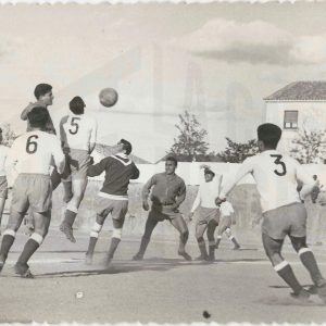 Varios futbolistas en una jugada durante un partido en el antiguo Campo de Fútbol de Aranjuez. Al fondo casas militares.