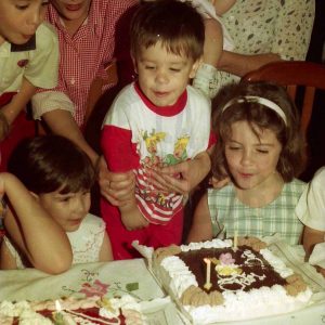 Un niño ante su tarta de cumpleaños con dos velas, rodeado de otros niños y niñas