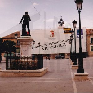 Cartel de «estamos rehabilitando ARANJUEZ» con vista posterior de la estatua de Alfonso XII en la Plaza de la Constitución en Aranjuez