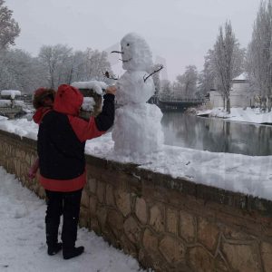 Una persona con un niño en brazos junto a un muñeco de nieve en el Jardín del Príncipe de Aranjuez al lado del río, al fondo el Puente Barcas