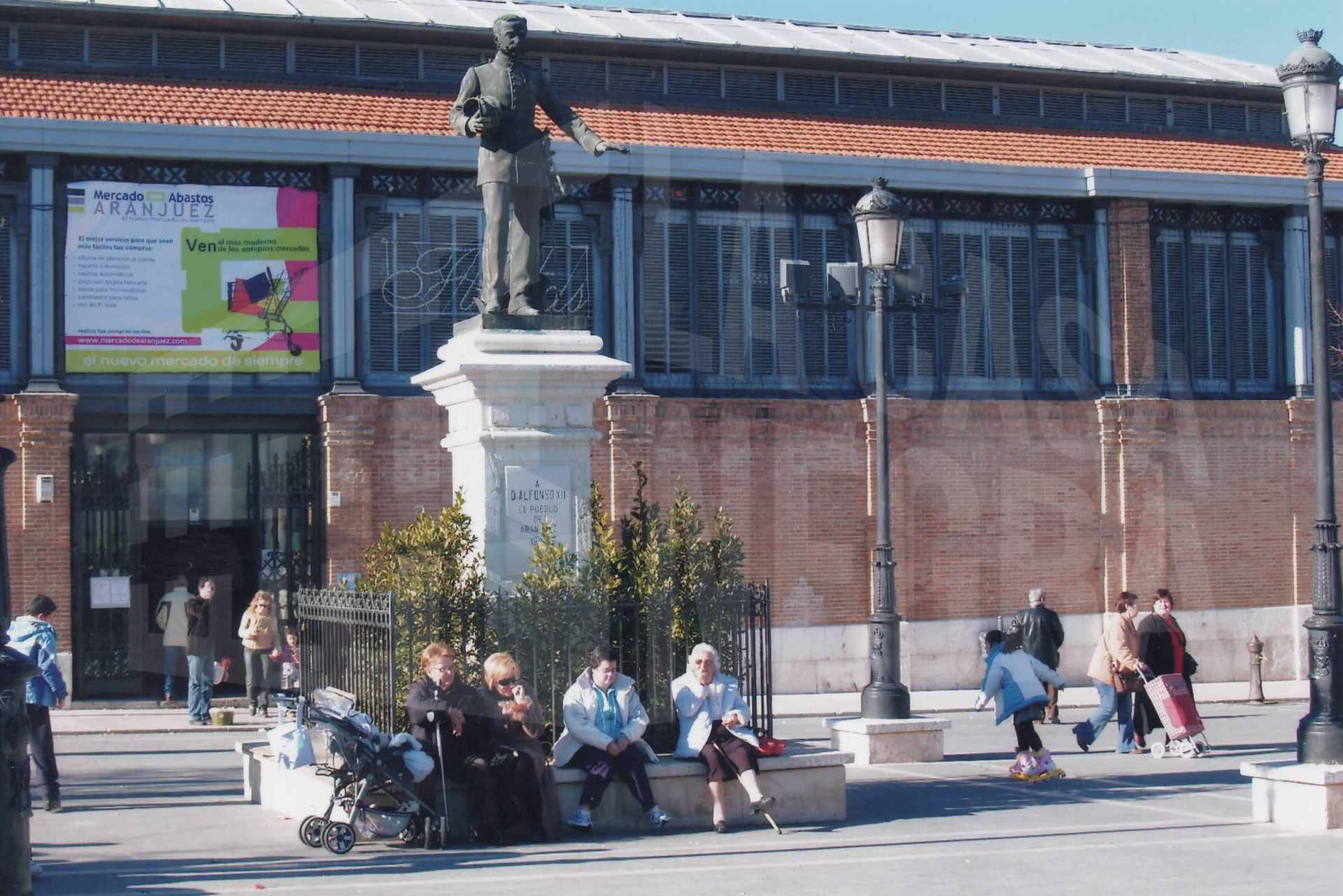 Varias personas alrededor de la puerta del Mercado de Abastos y de la estatua de Alfonso XII en la Plaza de la Constitución de Aranjuez